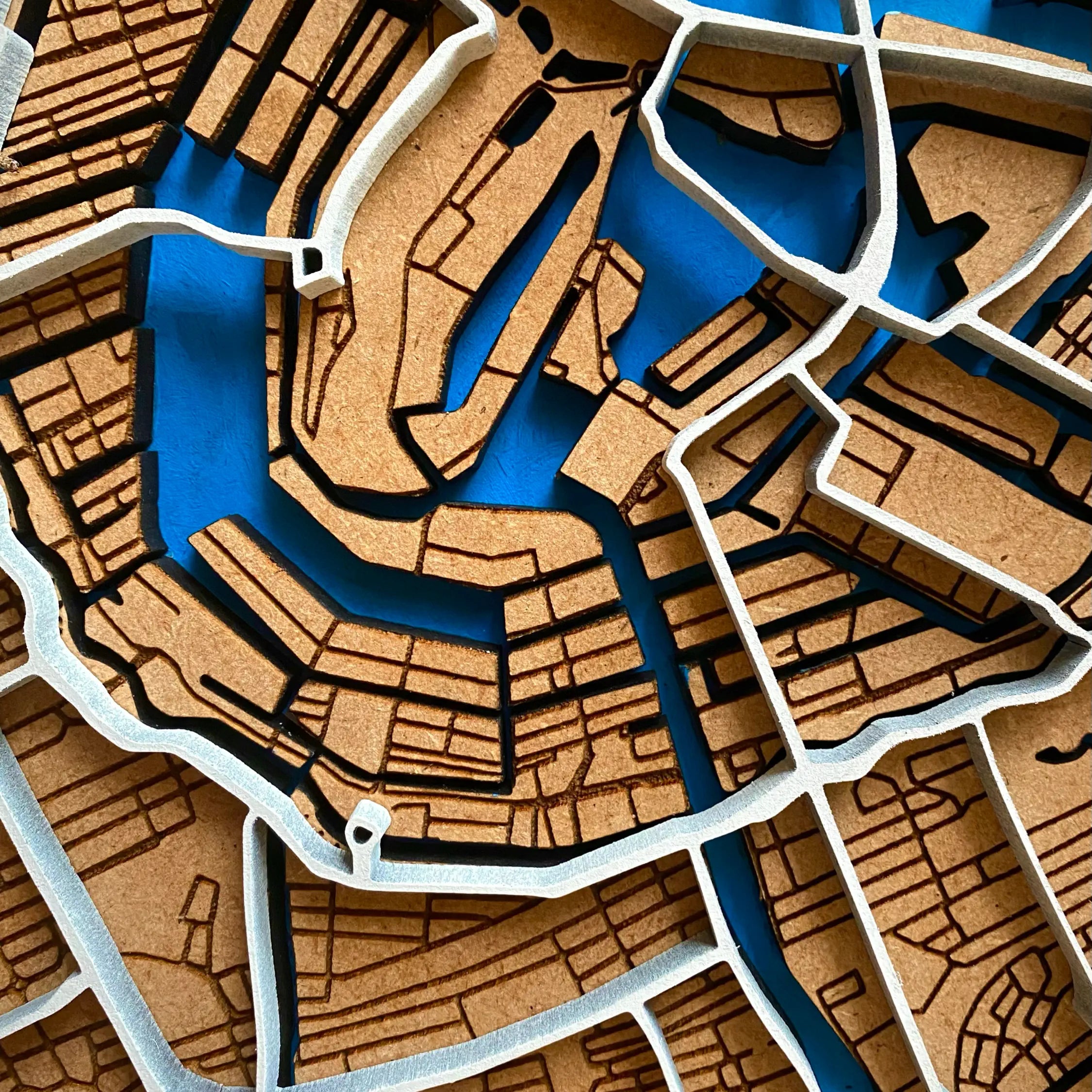 A super close-up view of the detailed wooden map with laser-cut small streets and blue hand-painted waterways. The top cut-out layer featured 3D elevated motoways and roundabouts which are hand-painted in white